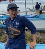 Seorang pegawai lapangan MDPI Bali bersandar di atas jukung. Namanya Putu Agus Widi, mengenakan topi, dan tampak bangga dengan seragam MDPI setelah mendampingi puluhan nelayan mendaftarkan kapalnya.